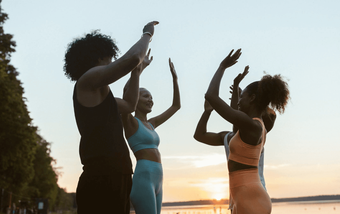 Group in activewear celebrating with raised hands by a lake, the sun low on the horizon